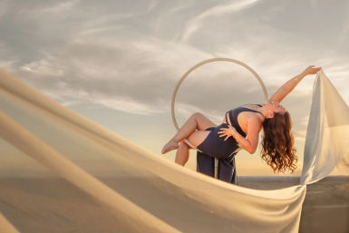 Graceful woman in a navy dress poses on a circular aerial hoop over desert dunes at sunset, billowing fabric and warm golden sky in an outdoor photoshoot.