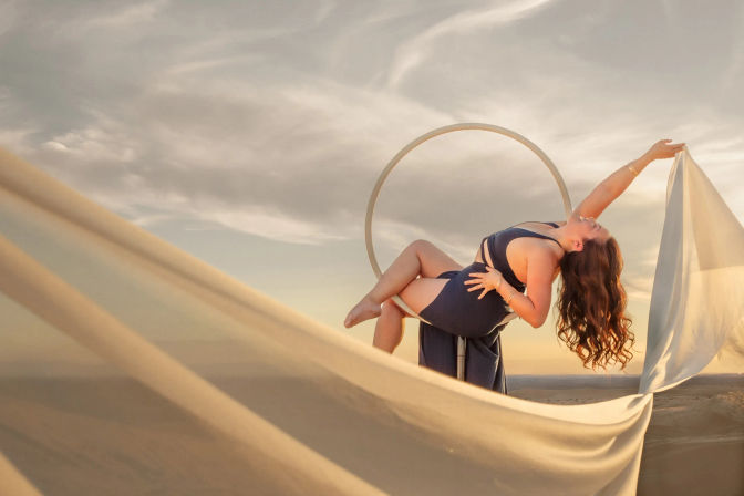Graceful woman in a navy dress poses on a circular aerial hoop over desert dunes at sunset, billowing fabric and warm golden sky in an outdoor photoshoot.