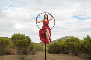 Woman in a flowing red dress perched on a black aerial hoop (lyra) above dry shrubland and rolling hills under a cloudy sky