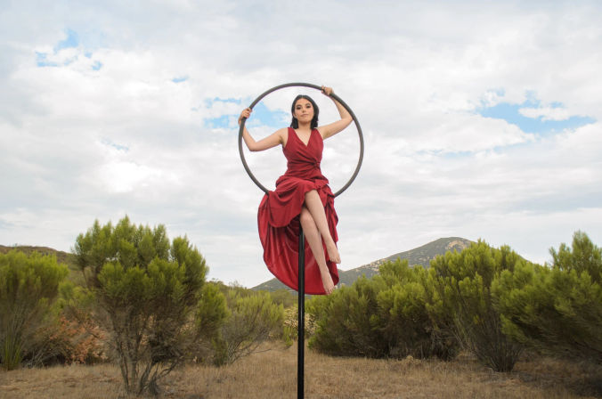 Woman in a flowing red dress perched on a black aerial hoop (lyra) above dry shrubland and rolling hills under a cloudy sky