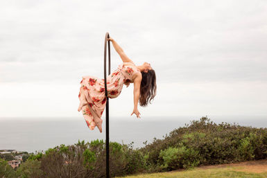 Aerial hoop performer in a flowing pink floral dress arches backward on a hoop above a coastal bluff with ocean horizon and cloudy sky