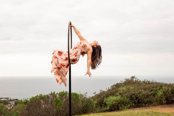 Aerial hoop performer in a flowing pink floral dress arches backward on a hoop above a coastal bluff with ocean horizon and cloudy sky