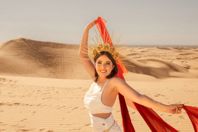 Smiling woman in a white dress wearing a golden rose sunburst crown and flowing red scarf, posing on sunlit sand dunes in a desert landscape.