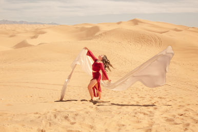 Joyful woman in a flowing red dress dancing with long white fabric across sunlit sand dunes, wide desert landscape of rolling golden dunes under a pale sky.
