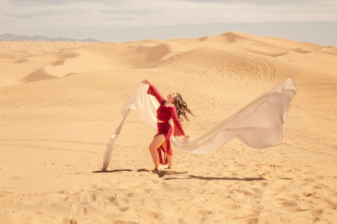 Joyful woman in a flowing red dress dancing with long white fabric across sunlit sand dunes, wide desert landscape of rolling golden dunes under a pale sky.