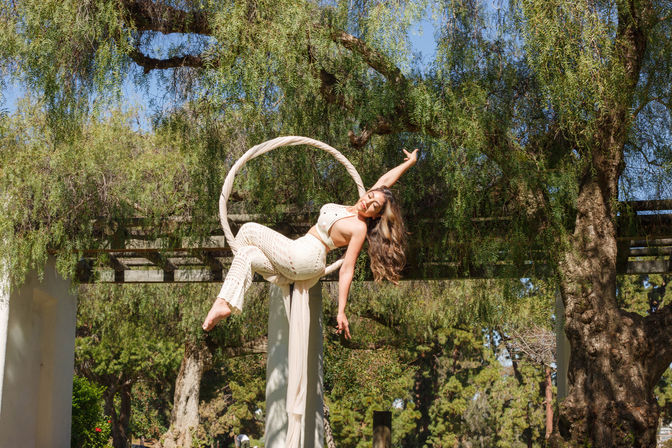 Aerialist on a fabric-wrapped hoop (lyra) in a cream crochet outfit, suspended under a sunlit pergola and stretching playfully among weeping willow trees in a leafy outdoor garden.