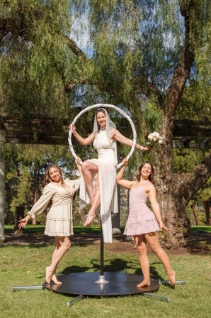 Playful outdoor wedding scene: a bride in a white dress and veil perched on a white aerial hoop holding a bouquet, flanked by two smiling friends in pastel dresses posing barefoot on a circular platform beneath hanging tree branches on a sunny day.
