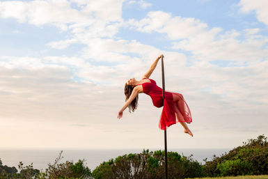 Pole dancer in a flowing red dress suspended on an outdoor pole, arched back and arms outstretched against a cloudy sky and seaside horizon with coastal shrubs