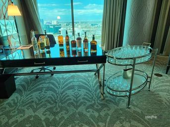High-rise hotel room bar setup: liquor bottles lined on a glossy desk beside a two-tier rolling cart stacked with glassware, with a floor-to-ceiling window showing a bright city skyline view.