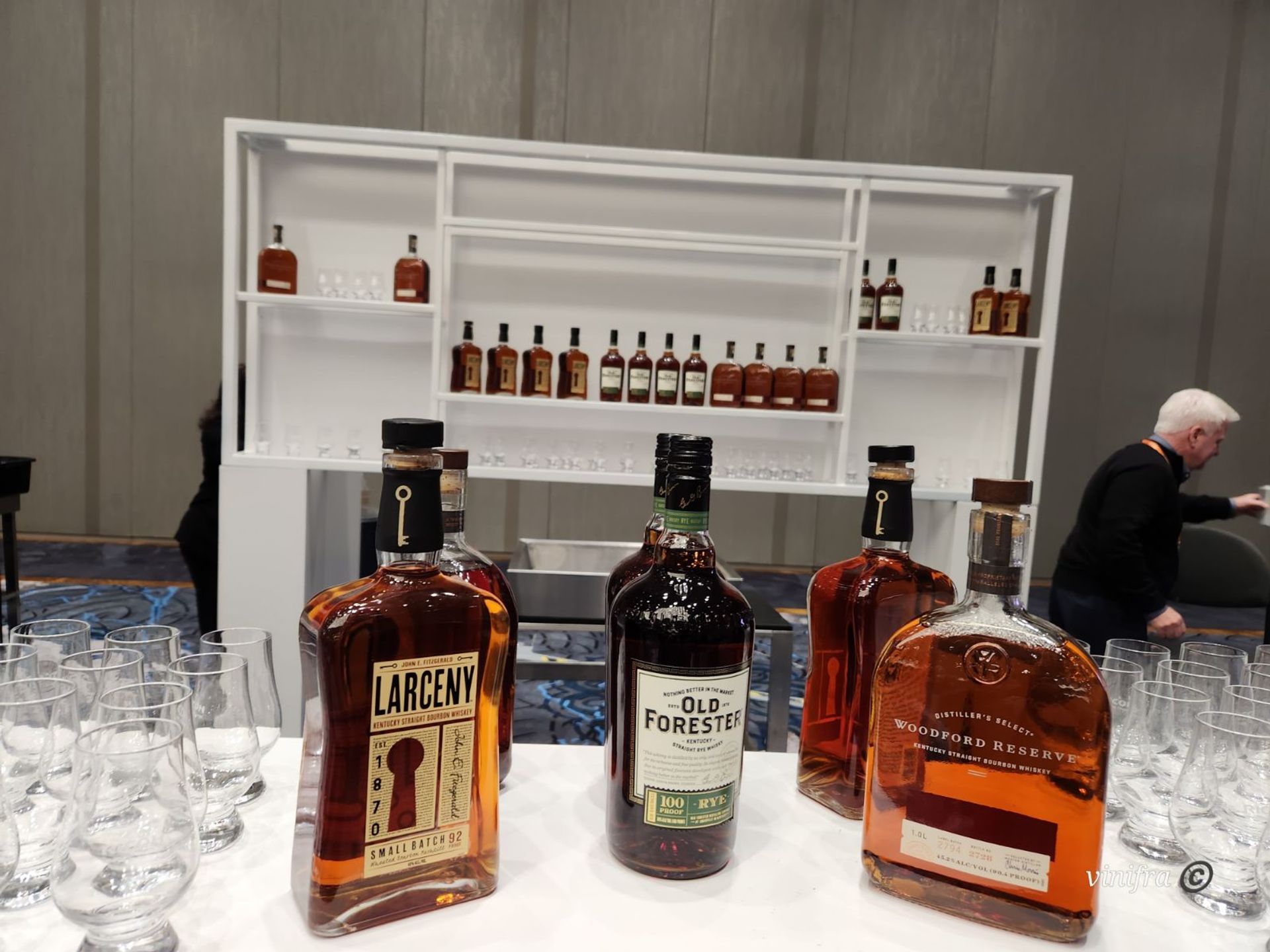 Bourbon tasting display with several whiskey bottles and rows of tasting glasses on a white table, bottle-filled shelving and conference room backdrop.