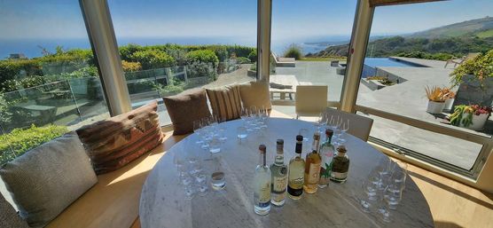 Sunlit coastal villa interior with a round marble table staged for a spirits tasting—bottles and multiple glasses on the table, cushioned banquette seating, panoramic glass walls opening to a patio with an infinity pool, coastal cliffs and ocean view.