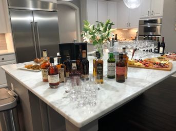 Modern white kitchen with a marble island set for a whiskey tasting — assorted bottles, tasting glasses, wine glasses, a charcuterie board with cheeses and cured meats, and a vase of white lilies.