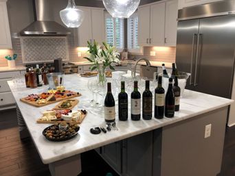 Bright modern kitchen with marble island staged for a wine tasting — row of wine bottles and glasses, ice bucket, charcuterie boards and olives, vase of lilies, pendant lights and stainless-steel appliances.