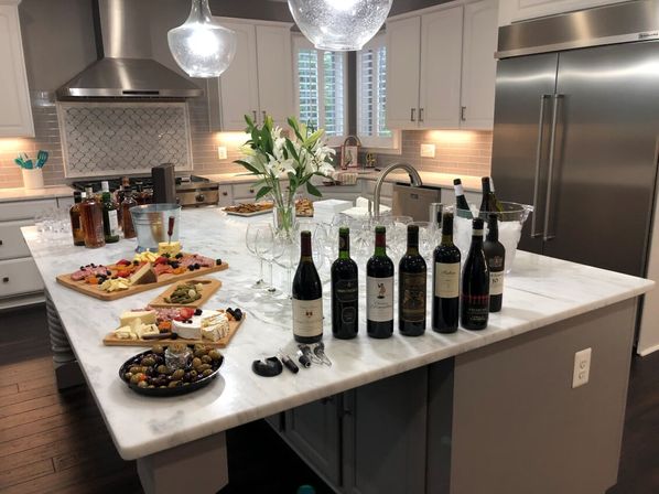 Bright modern kitchen with marble island staged for a wine tasting — row of wine bottles and glasses, ice bucket, charcuterie boards and olives, vase of lilies, pendant lights and stainless-steel appliances.