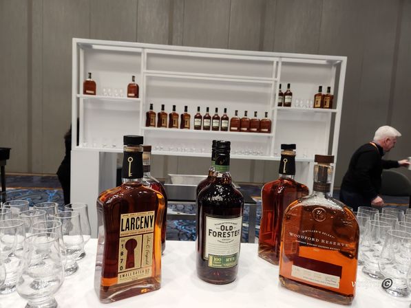 Whiskey tasting display with Larceny, Old Forester and Woodford Reserve bottles on a table, empty tasting glasses and a white shelving unit of bottles in an indoor event room.