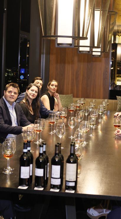 Wine tasting in an upscale high-rise private dining room at night, guests seated along a long table with glasses and bottles as a presenter speaks