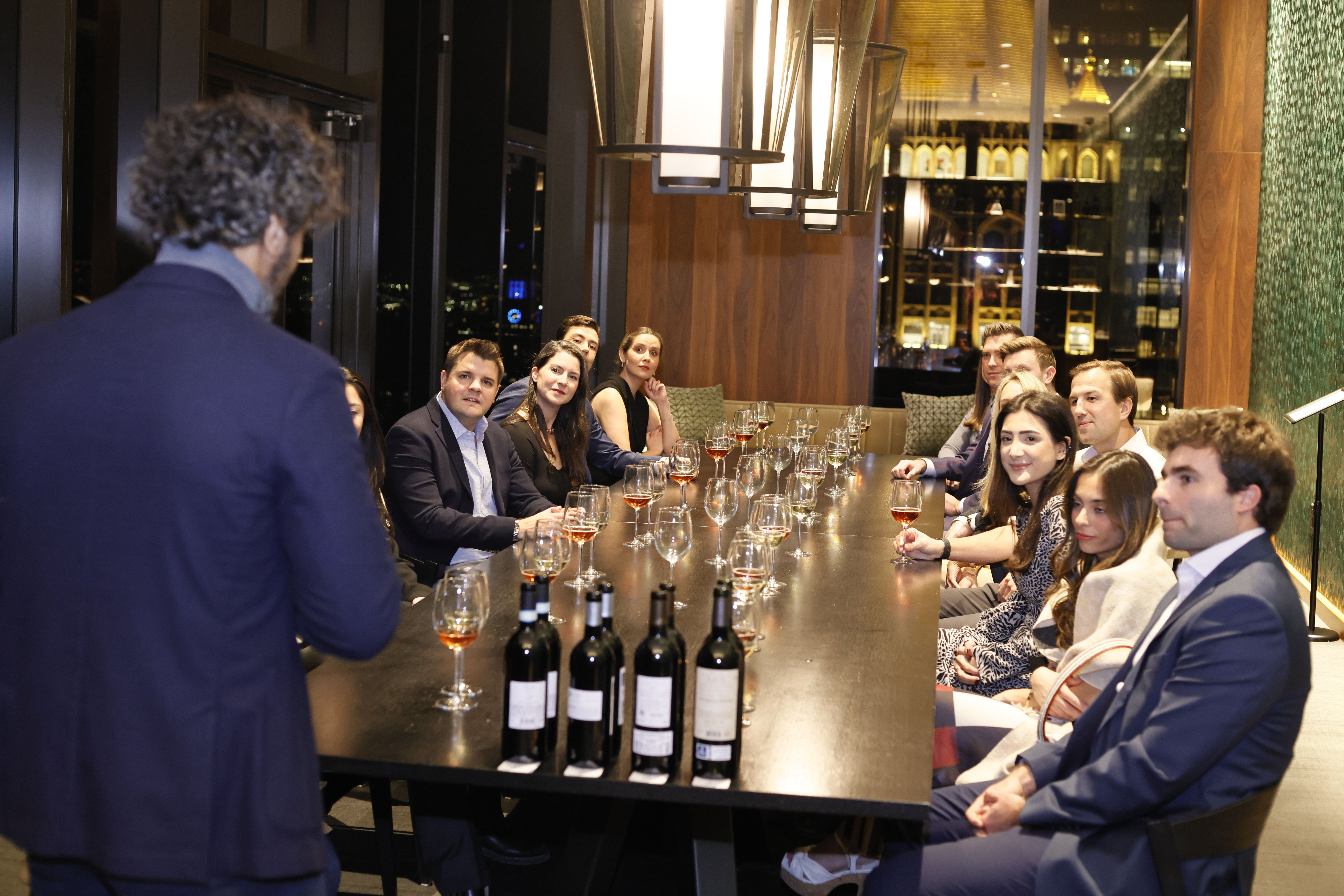 Private wine tasting in a chic high‑rise dining room with city skyline views, guests seated at a long table listening to a host with wine glasses and bottles on display.