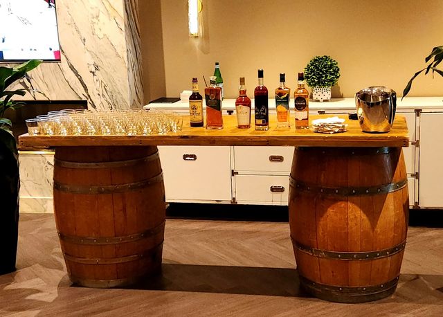Rustic whiskey tasting setup on a plank table supported by two oak barrels, with rows of tasting glasses, assorted bottles, ice bucket and napkins in an indoor event space.