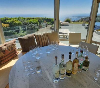 Coastal oceanfront dining room with round marble table set for spirits tasting—bottles and multiple tasting glasses, cushioned banquette, panoramic windows, glass railing, infinity pool and rugged sea cliffs beyond.