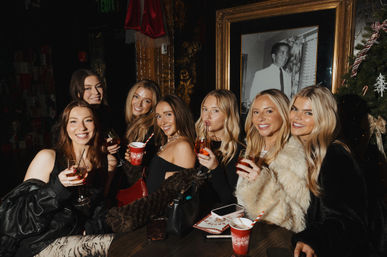 Seven smiling women toast with holiday cocktails and red festive cups at an indoor Christmas-themed party beside a decorated tree and framed portrait.