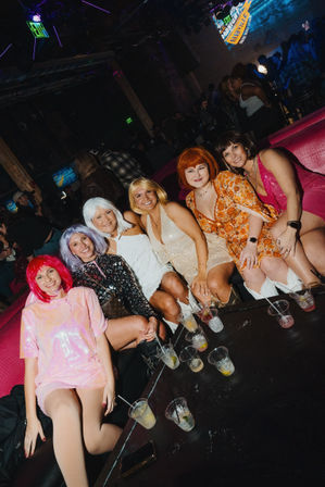 Group of six women in colorful wigs and vintage-style dresses smiling on a pink booth inside a lively nightclub lounge, cocktails on a low table in front of them