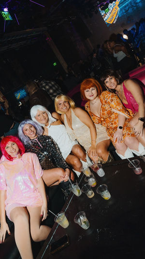 Group of six women in colorful wigs and vintage-style dresses smiling on a pink booth inside a lively nightclub lounge, cocktails on a low table in front of them