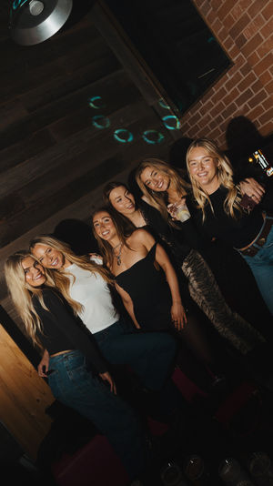 Six friends smiling and posing for a night out at a dim indoor bar lounge with exposed brick and wood walls, disco light reflections, casual outfits and drinks