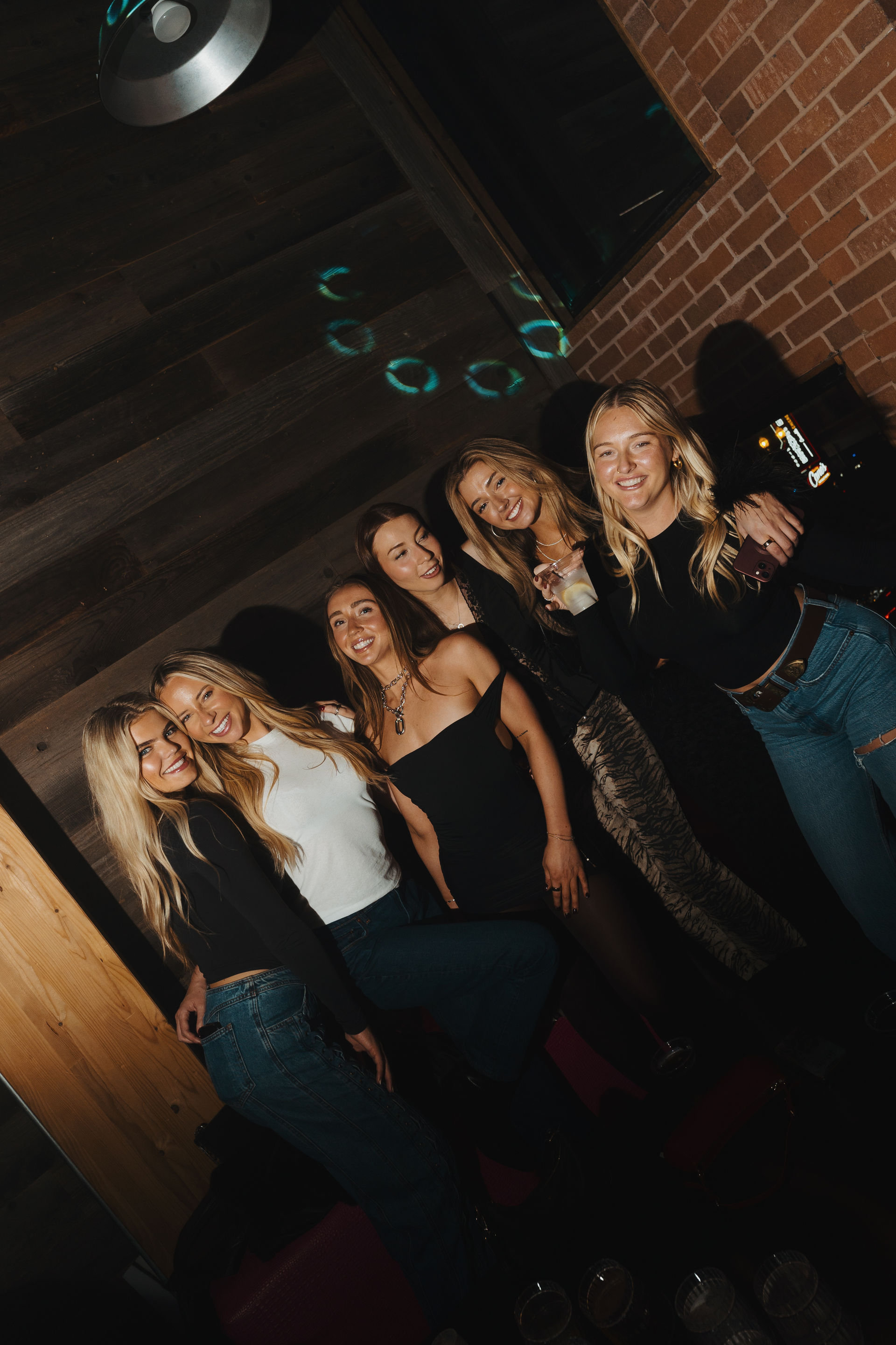 Six friends smiling and posing for a night out at a dim indoor bar lounge with exposed brick and wood walls, disco light reflections, casual outfits and drinks