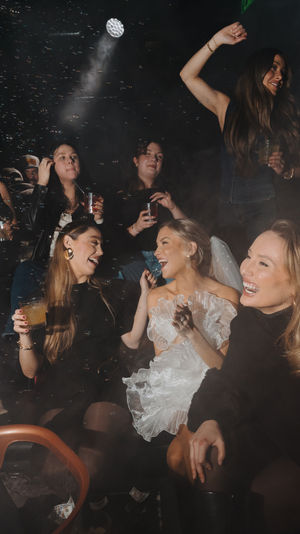 Lively nightclub bachelorette party with friends laughing, holding cocktails, and a bride-to-be in a ruffled white dress and veil under smokey disco lights