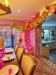 Home dining room bachelorette party setup with pink balloon arch, gold foil letter balloons spelling BRIDE, shimmering pink fringe backdrop, table set with pink plates, cups and festive place settings, and a view into a modern kitchen.