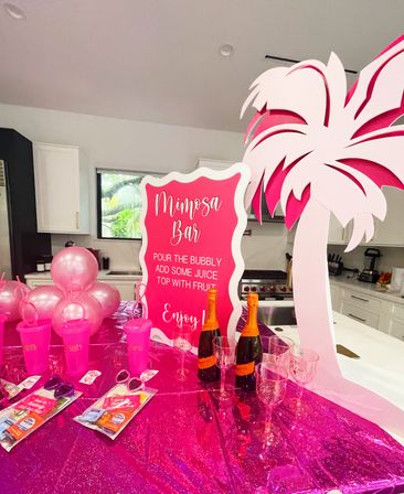 Bright pink mimosa bar on a home kitchen island with a glittery fuchsia tablecloth, metallic pink balloons, pink plastic cups and party favors, two bottles of sparkling wine, clear glasses, a large decorative pink palm cutout and a bold pink sign reading "Mimosa Bar" with pour-and-top instructions.