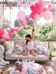 Bachelorette party on a backyard patio swing with pink balloon garland, mirrored disco balls, and a woman in a bride swimsuit raising a champagne bottle