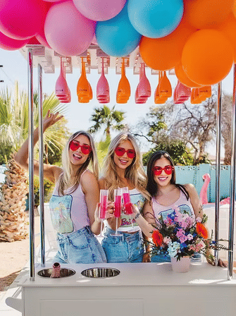 Three friends in red heart sunglasses toast pink cocktails at a colorful balloon-topped poolside cart with palm trees and flamingo decor, summer party vibe
