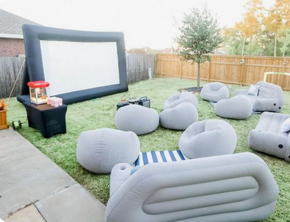 Cozy backyard outdoor movie night setup on a green lawn with a large inflatable projection screen, gray inflatable sofas and beanbag chairs, a popcorn machine on a side table, and a wooden privacy fence