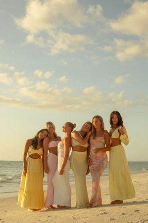 Six women in pastel summer dresses posing together on a sandy beach at golden hour with a calm ocean and soft clouds — coastal sunset group portrait