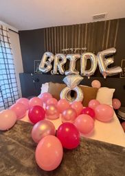 Cozy bedroom bridal surprise with silver foil 'BRIDE' balloons and oversized diamond ring balloon over bed, pink and rose-gold balloons scattered across white bedding and dark wood accent wall.
