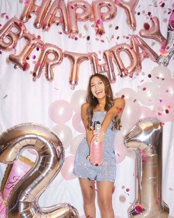 Smiling woman celebrating her 21st birthday indoors with rose-gold "HAPPY BIRTHDAY" banner, giant rose-gold "21" balloons, pink and white balloons, confetti, and a sparkling mini dress while holding a confetti popper.