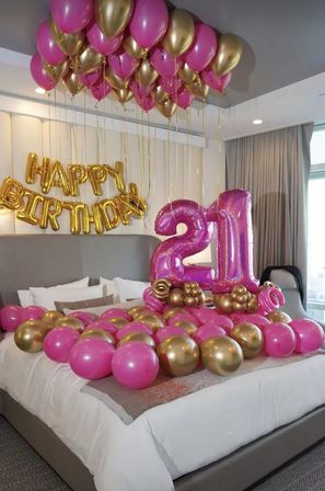 Festive bedroom setup with pink and gold balloons tied to the ceiling, giant pink foil ‘21’ numbers on the bed, and a gold ‘Happy Birthday’ foil banner on the wall