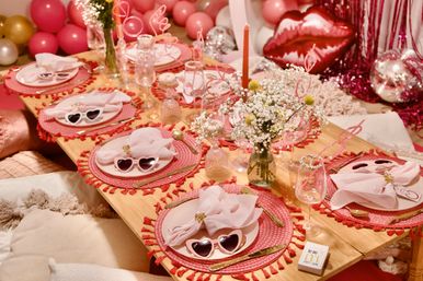 Pink festive low table set for a party with heart-shaped sunglasses on blush plates, tasseled placemats, gold cutlery, sheer napkins, baby's-breath centerpieces and pink and gold balloons