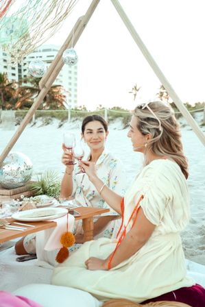 Boho beach picnic — two women clinking wine glasses under a wooden teepee decorated with disco balls on a sandy oceanfront beach with palm trees and a hotel in the background.