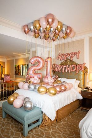 Festive hotel bedroom decorated for a 21st birthday with rose-gold, gold, and silver balloons clustered on the ceiling, large foil '21' numbers and 'Happy Birthday' balloons above a made bed.