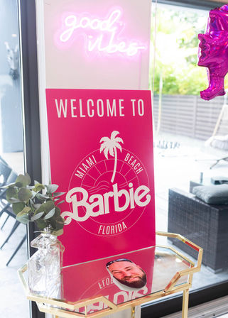 Sunlit interior with bright pink Miami Beach, Florida–themed sign on a mirrored gold table, neon "good vibes" glow overhead, glass vase of greenery and a metallic pink balloon by a patio door.
