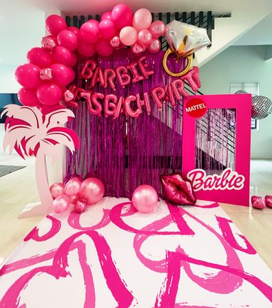 Vibrant pink indoor party photo booth with shimmering fringe backdrop, balloon garland and arch, oversized diamond‑ring and lips balloons, palm tree cutout, pink frame prop, and bold heart‑brushstroke floor runner.