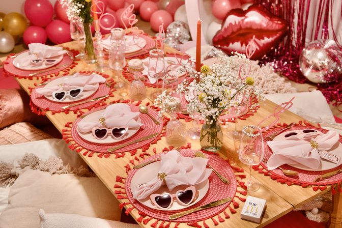 Pink Valentine's party tablescape with heart-shaped sunglasses, tassel placemats, gold cutlery, folded napkins, baby's-breath bouquets and balloons