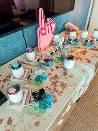 Hotel suite welcome table set for a bachelorette party with a pink foam hand prop, white insulated tumblers, pink cat-eye sunglasses, rose-gold ring confetti and packaged party favors on a marble countertop.