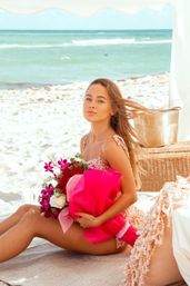 Sunlit seaside scene: woman seated on a sandy beach under a white canopy, holding a bright pink-wrapped bouquet next to a wicker picnic basket with turquoise ocean waves in the background