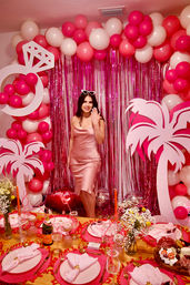 Glam indoor pink celebration: woman in a pink satin dress showing a ring in front of a pink balloon arch and metallic fringe backdrop, palm cutouts and a pink-themed table set with champagne and place settings.