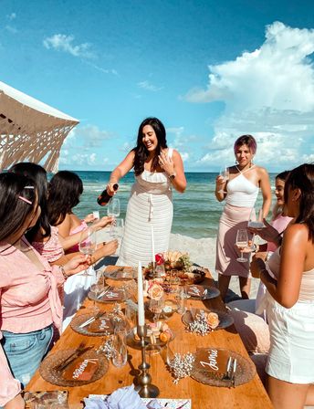 Seaside champagne toast with friends at a boho beach picnic: wooden table set with candles, roses, charcuterie and an ocean backdrop