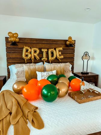 Cozy bridal bedroom setup with gold foil 'BRIDE' balloons on a wooden headboard, green, orange and gold balloons scattered on a white quilted bed, mustard knit throw and a wooden tray with wine and two glasses.