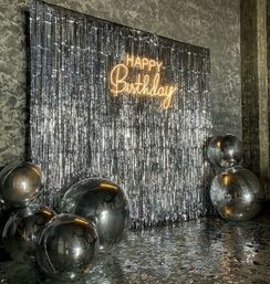 Silver metallic fringe photo backdrop with illuminated neon "Happy Birthday" sign and oversized reflective silver balloons on the floor, glittering indoor party photo-op setup.